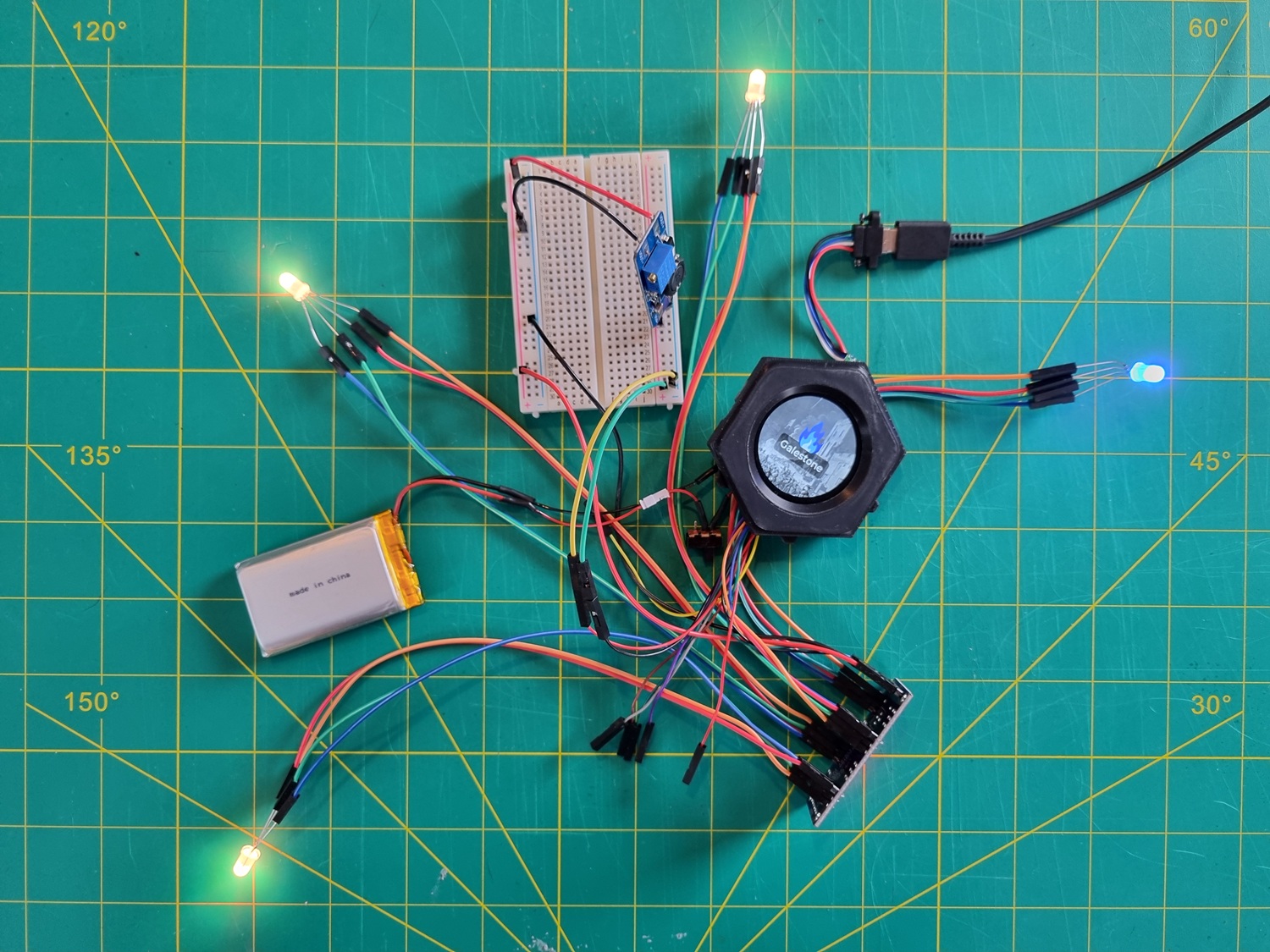 The electronics prototype laid out on a cutting mat: a breadboard, the TLC5947 module, an MT3608 boost converter, the ESP32-S3 with its circular display, and a LiPo battery, wired together with Dupont connectors. The four RGB LEDs are lit and the display shows 'Galestone' as a blue flame, this colour reflected in the rightmost LED.