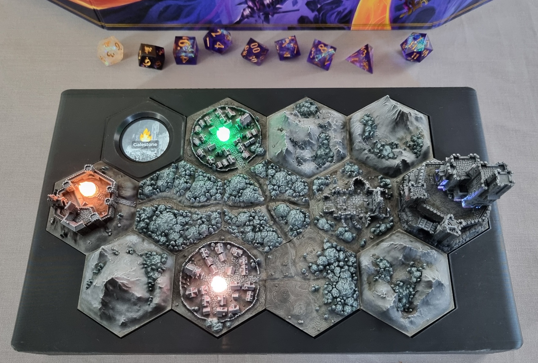 Photo of the ESP32-powered interactive TTRPG hex map console showing black box housing a 3-row (4-5-4) hex tile grid, painted to look like moonlight. The ESP32-powered display is in the top left. LED fires flicker in each of the town tiles. A set of Daggerheart dice and the Daggerheart GM screen are visible above the console.