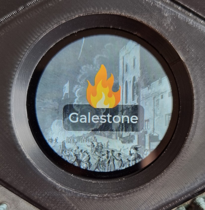 The circular display, embedded in its hex tile, showing the Galestone screen: a healthy orange flame icon atop a black-and-white painting of a town square. The circular display, embedded in its hex tile, showing the Galestone screen: a healthy orange flame icon atop a black-and-white painting of a town square.